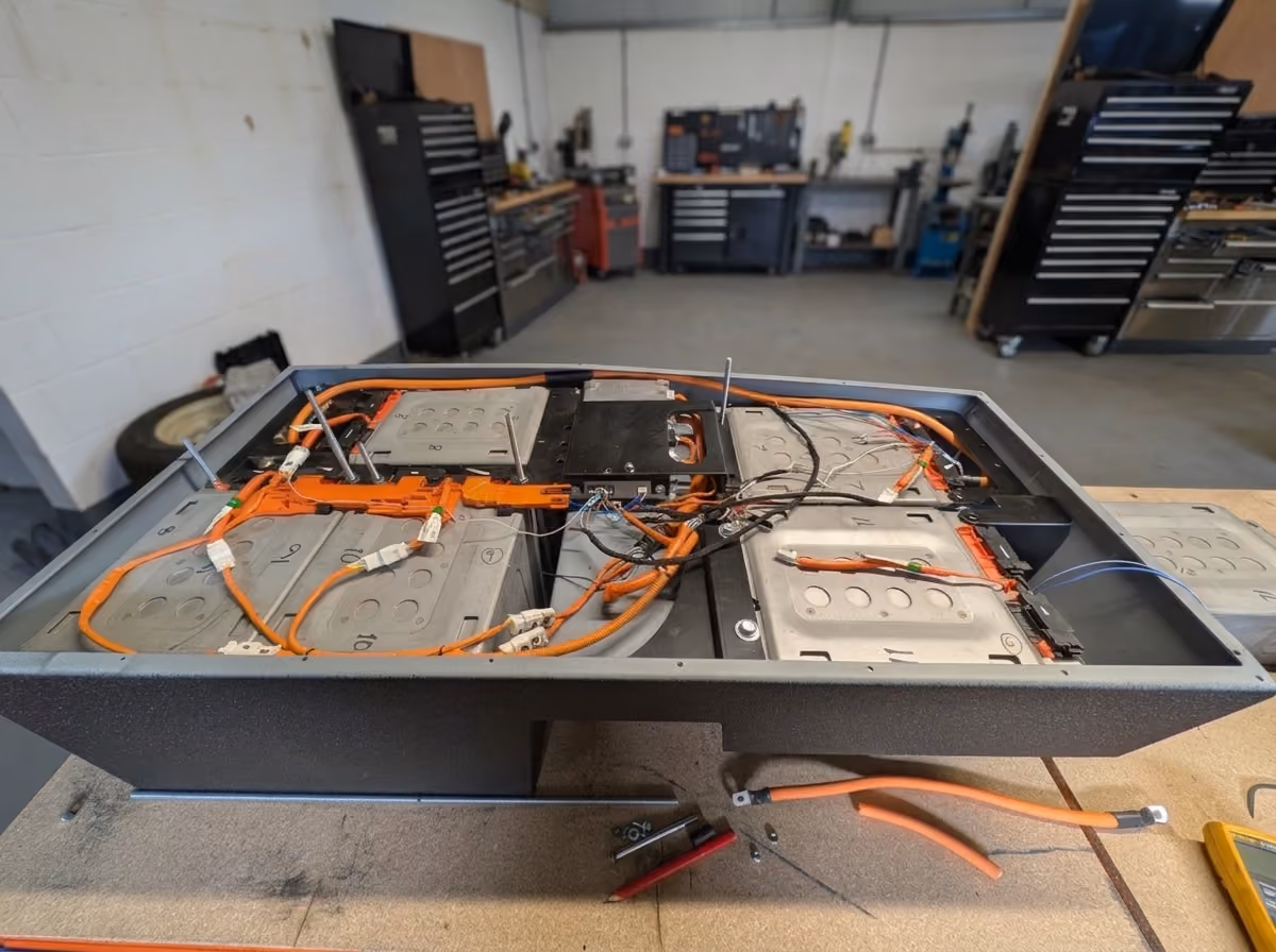 Open battery pack with multiple rectangular battery cells connected by orange high-voltage cables and black wires, placed on a workbench in a workshop with tool cabinets and equipment in the background.