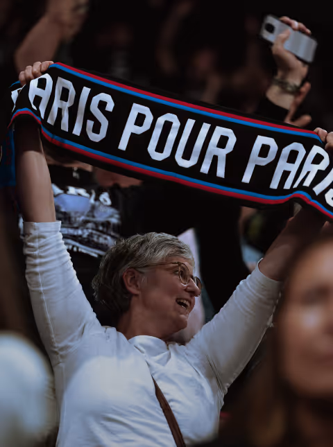 Woman in white shirt holding up a scarf that reads 'PARIS POUR PARA' at a crowded event.