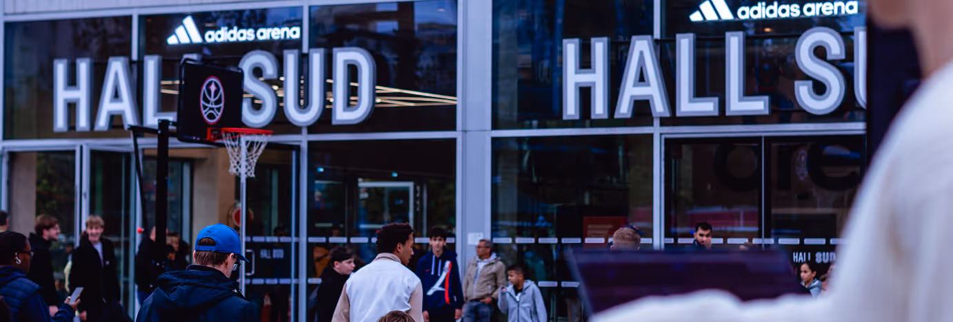 People gathered outside adidas arena near a basketball hoop and glass doors displaying 'HALL SUD'.