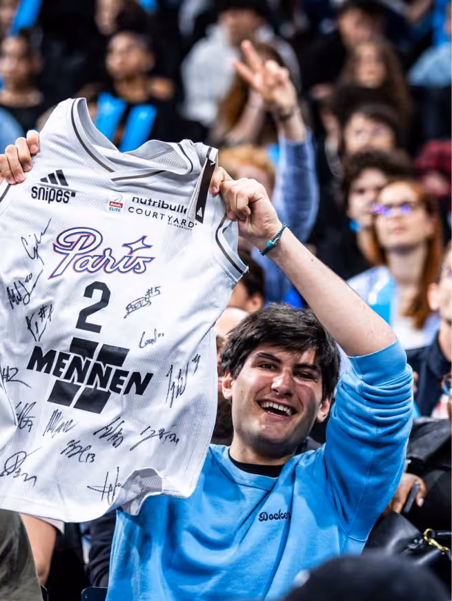 Smiling man in a blue shirt holding up a signed Paris basketball jersey with the number 2 at a crowded event.