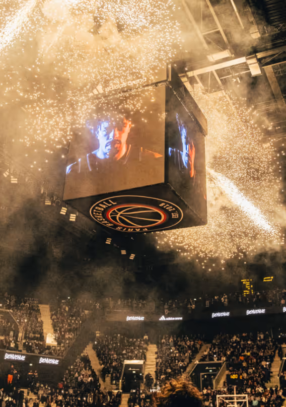 Basketball arena scoreboard displaying a player’s face with fireworks and smoke surrounding it above a crowd.
