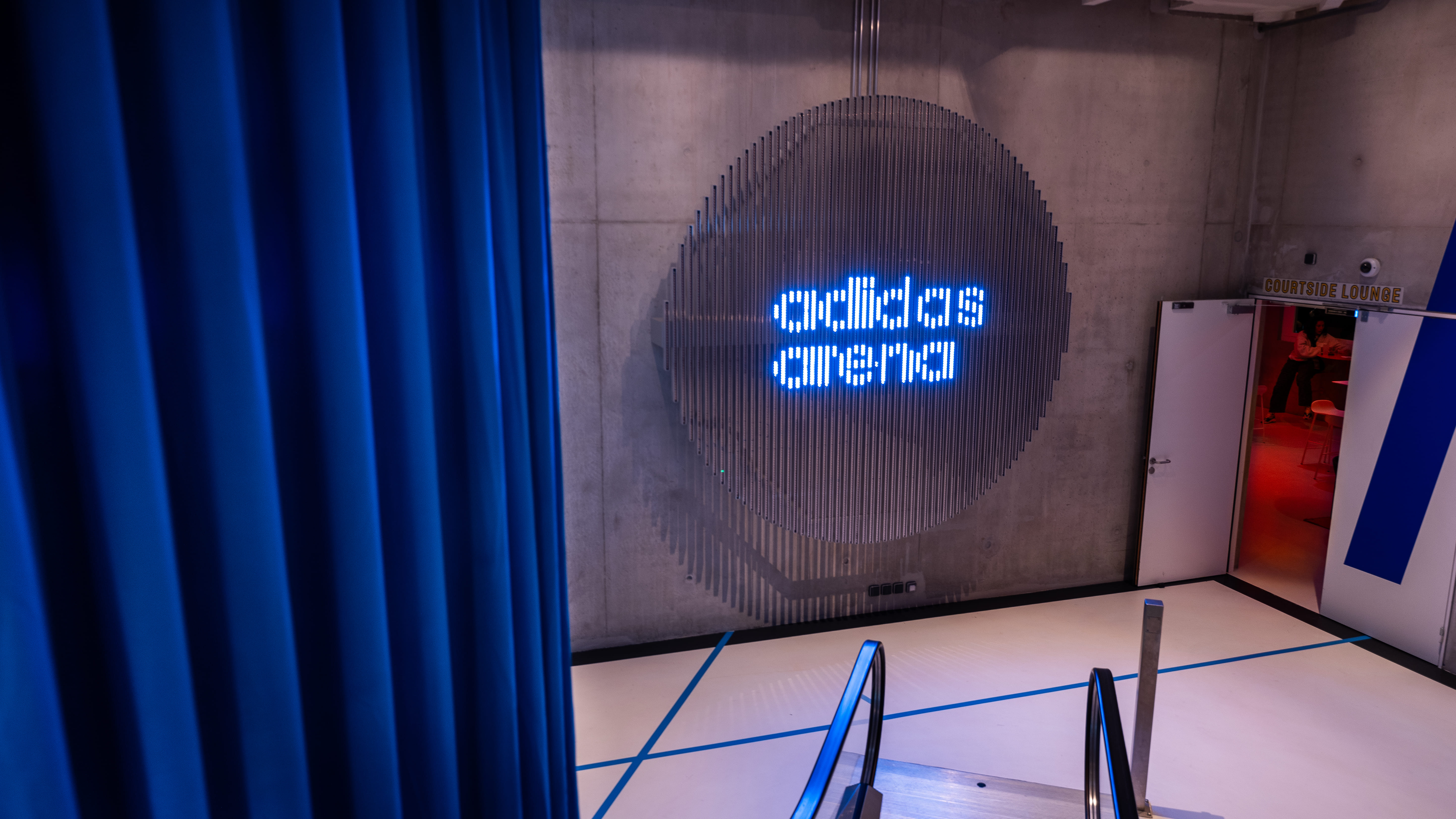 View down an escalator towards a wall with a blue illuminated adidas arena sign and an open door labeled Courtside Lounge with red lighting inside.