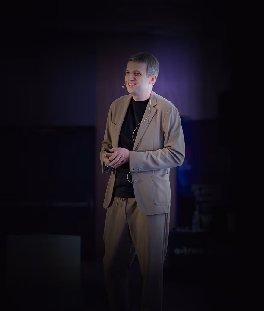Man smiling and speaking on stage wearing a headset microphone, beige suit, and black shirt against dark background.