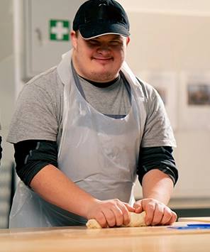 Smiling worker wearing a black cap and apron kneading dough in a professional kitchen.