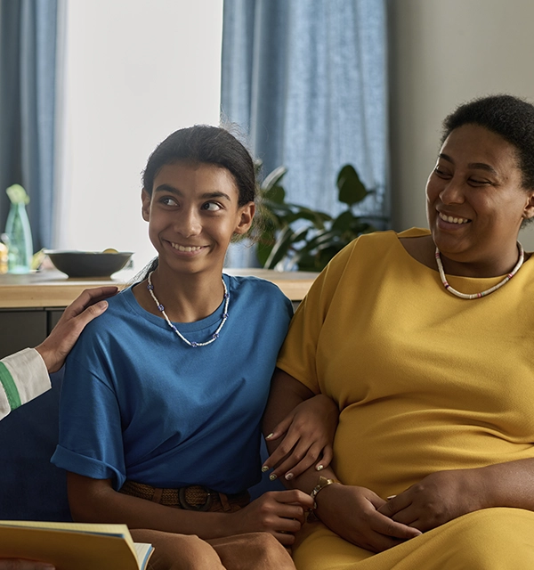 A smiling teenage girl in a blue shirt sitting next to a woman in a yellow dress who has her arm around the girl.