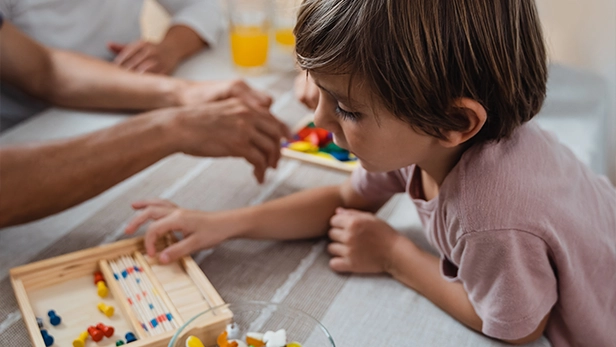 Young boy playing a wooden board game at a table with an adult's hands nearby.