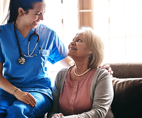 Smiling nurse in blue scrubs with stethoscope comforting elderly woman sitting on a couch.