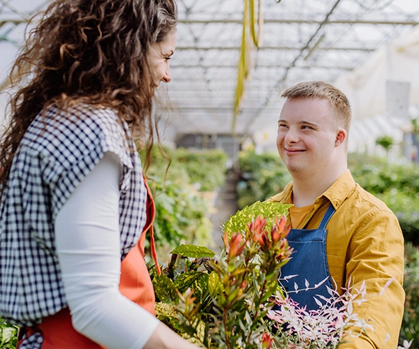 Two people smiling and holding plants inside a greenhouse, surrounded by greenery.