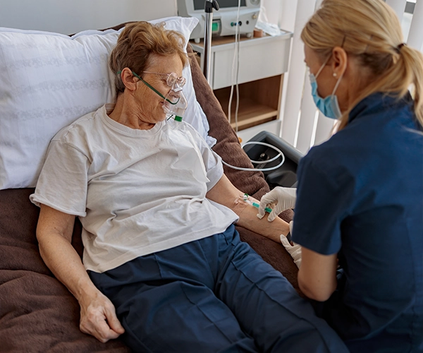 Nurse in blue scrubs and mask administering an intravenous line to a senior patient lying in a hospital bed with oxygen nasal cannula.