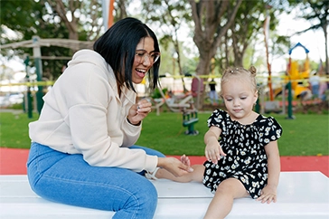 Woman in a white hoodie engaging and playing with a young girl in a black floral dress at a park.