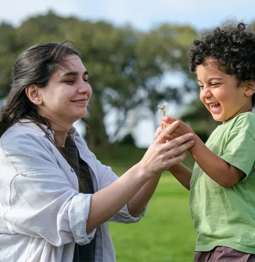 Woman and young boy smiling and holding a dandelion together outdoors in a green park.