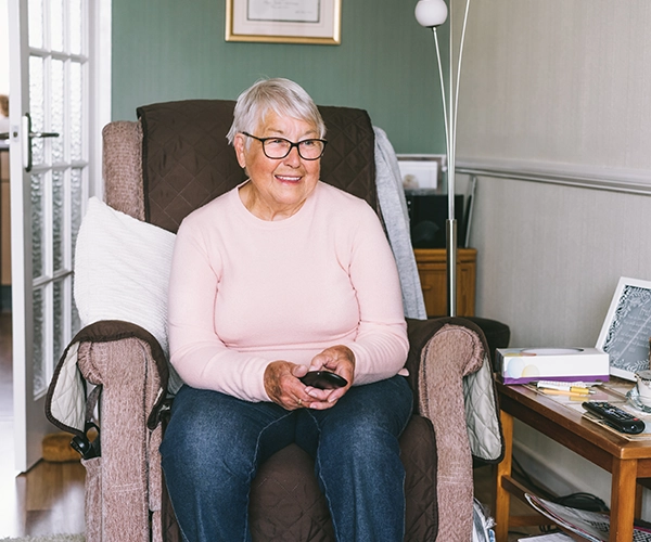 Elderly woman with glasses sitting in a cozy armchair holding a TV remote, smiling in a living room.