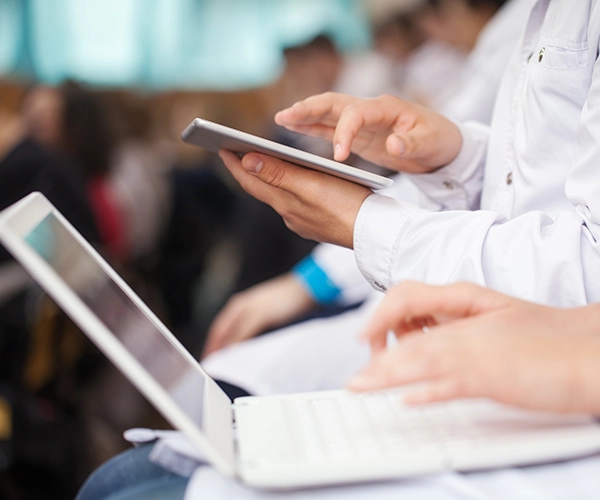 Medical students in white coats using a tablet and a laptop side by side.