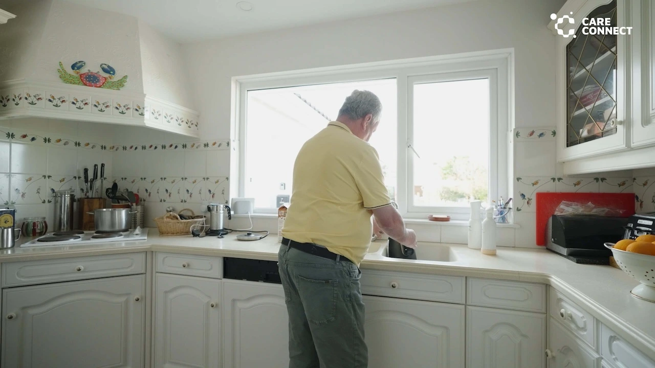An elderly man in a yellow shirt washing dishes at a kitchen sink under a window.