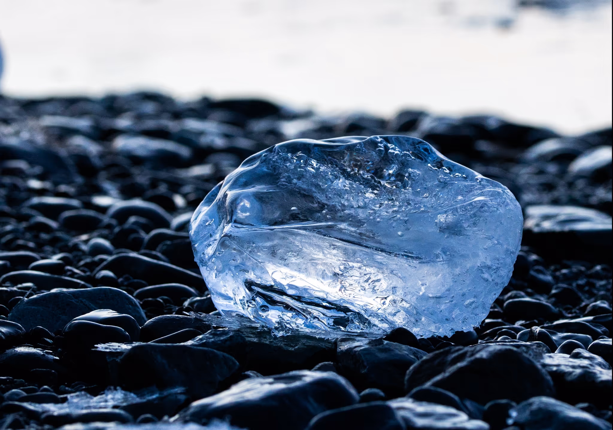 Ein glatter, transparenter Eisblock liegt auf dunklen, nassen Steinen am Ufer.