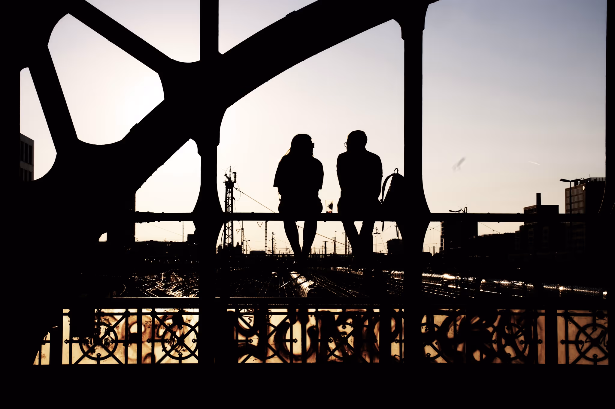 Silhouetten eines Mannes und einer Frau, die auf einem Geländer über einem Bahngleis sitzen und beim Sonnenuntergang sprechen.