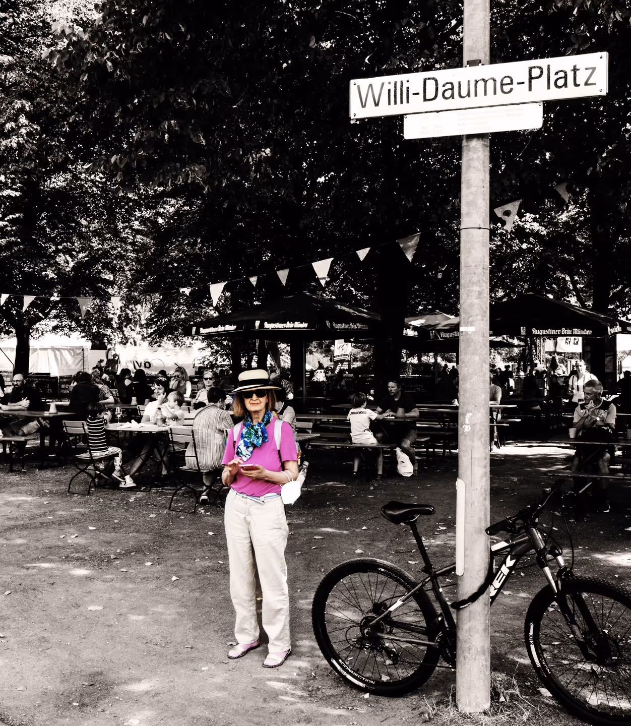 Frau mit Hut, violettem Shirt und weißer Hose steht vor einem Fahrrad an einem Pfosten mit Straßenschild Willi-Daume-Platz, im Hintergrund Menschen an Biertischen unter Bäumen.