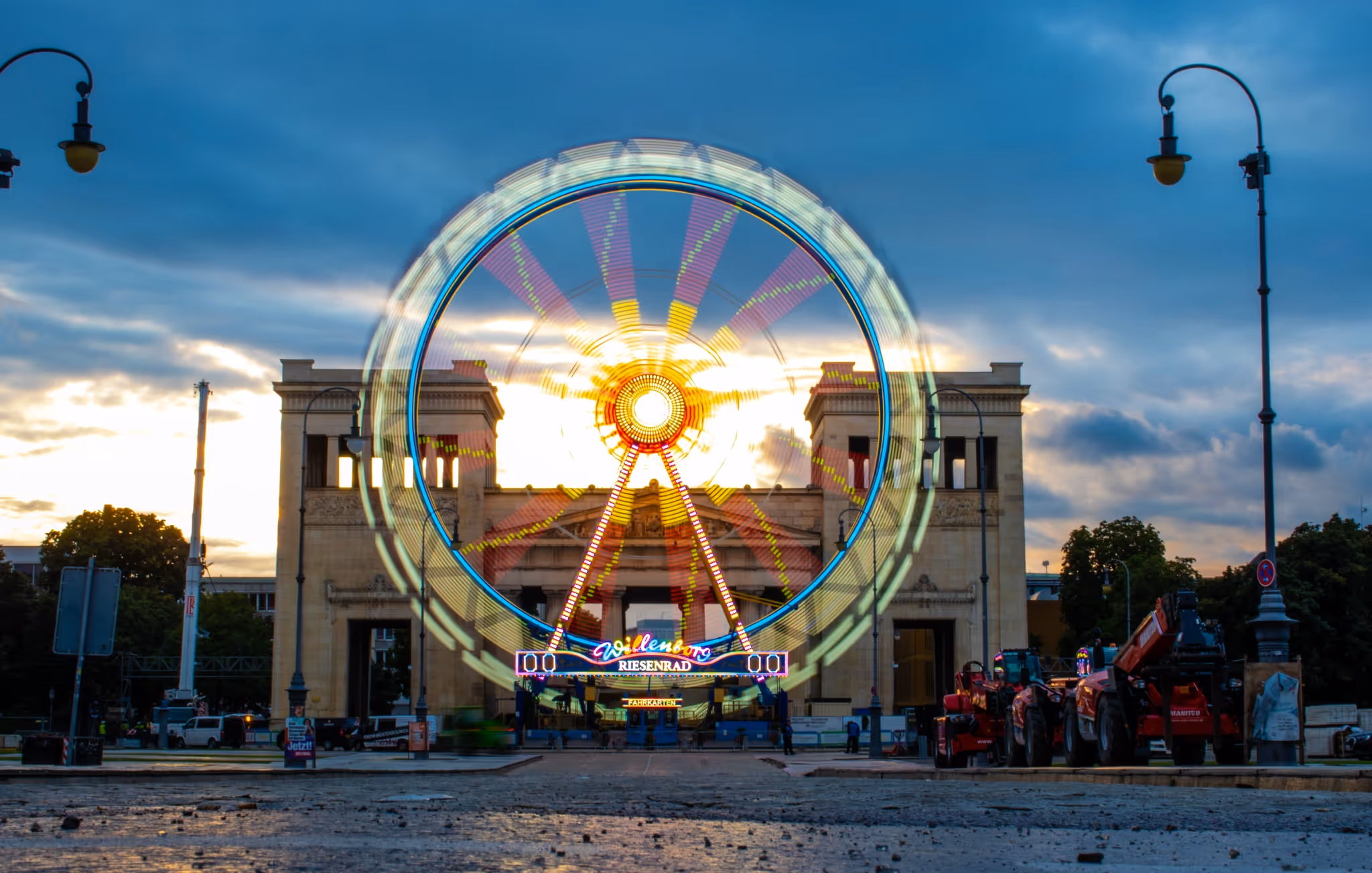 Beleuchtetes Riesenrad mit Bewegungseffekt vor einem historischen Gebäude bei Dämmerung.