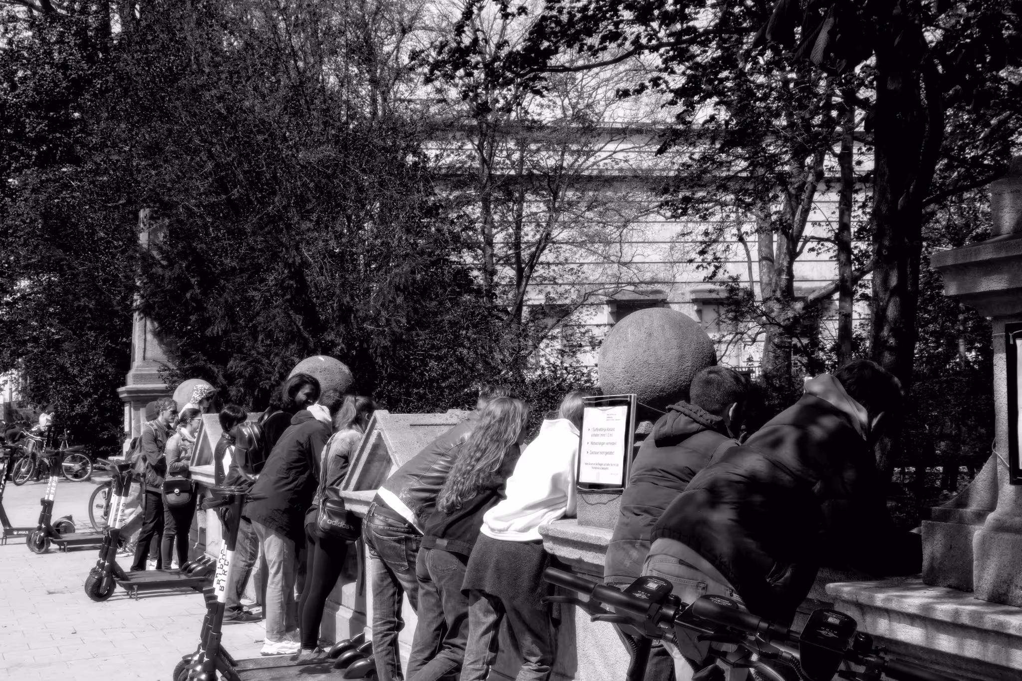 Mehrere Menschen stehen an einer Steinmauer und schauen in Informationstafeln, im Hintergrund Bäume und Roller.