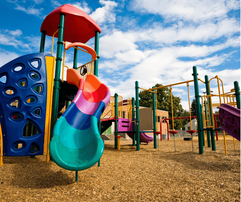 A vibrant children's playground under a clear blue sky, with multi-colored slides, blue and purple climbing frames, red swings, and yellow and green bars, set on a wood chip surface with trees and a red brick building in the background.