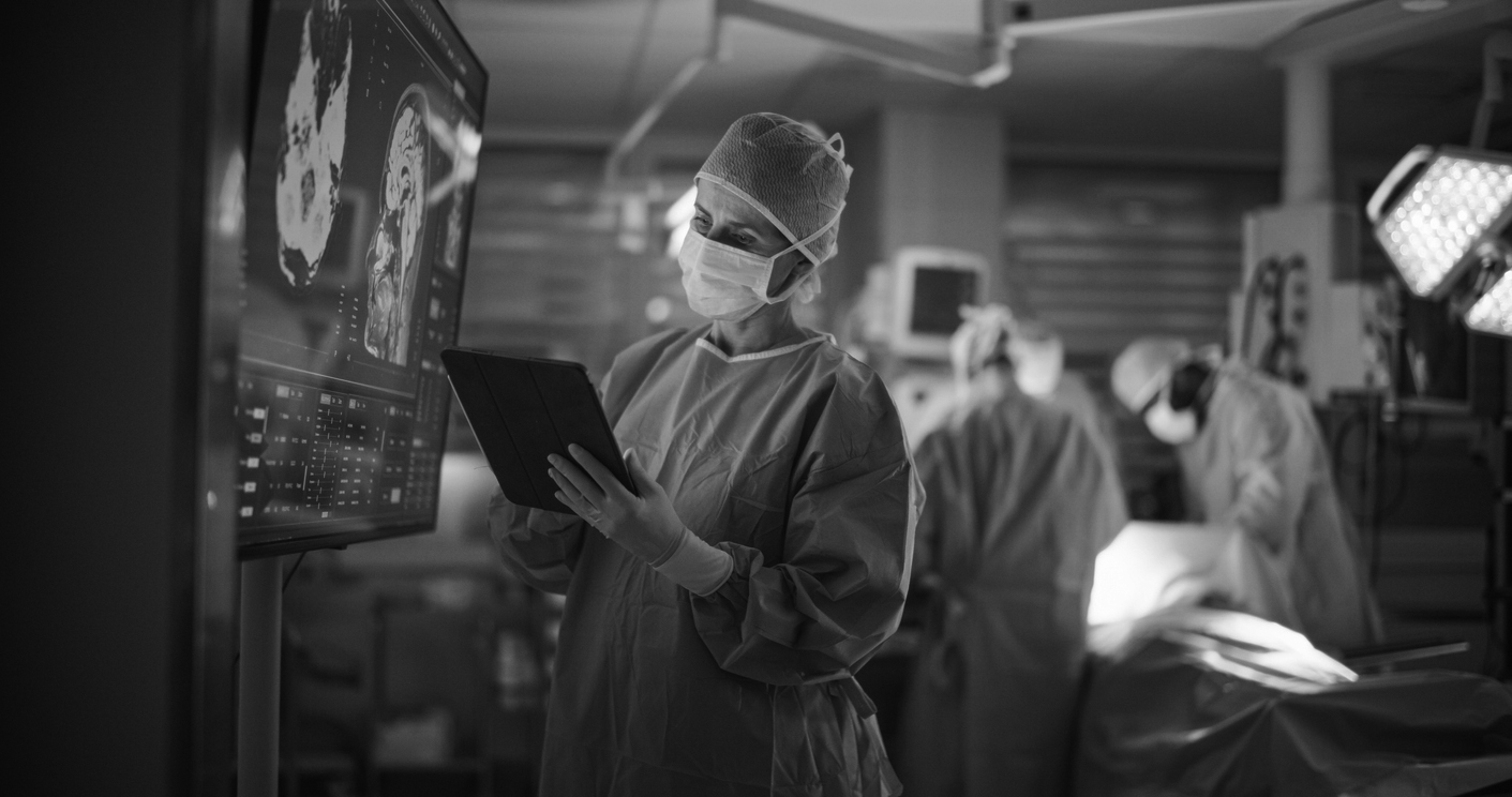 A nurse in the foreground is reviewing a patients chart in the operating room while two other nurses in the background are assisting with an operation.