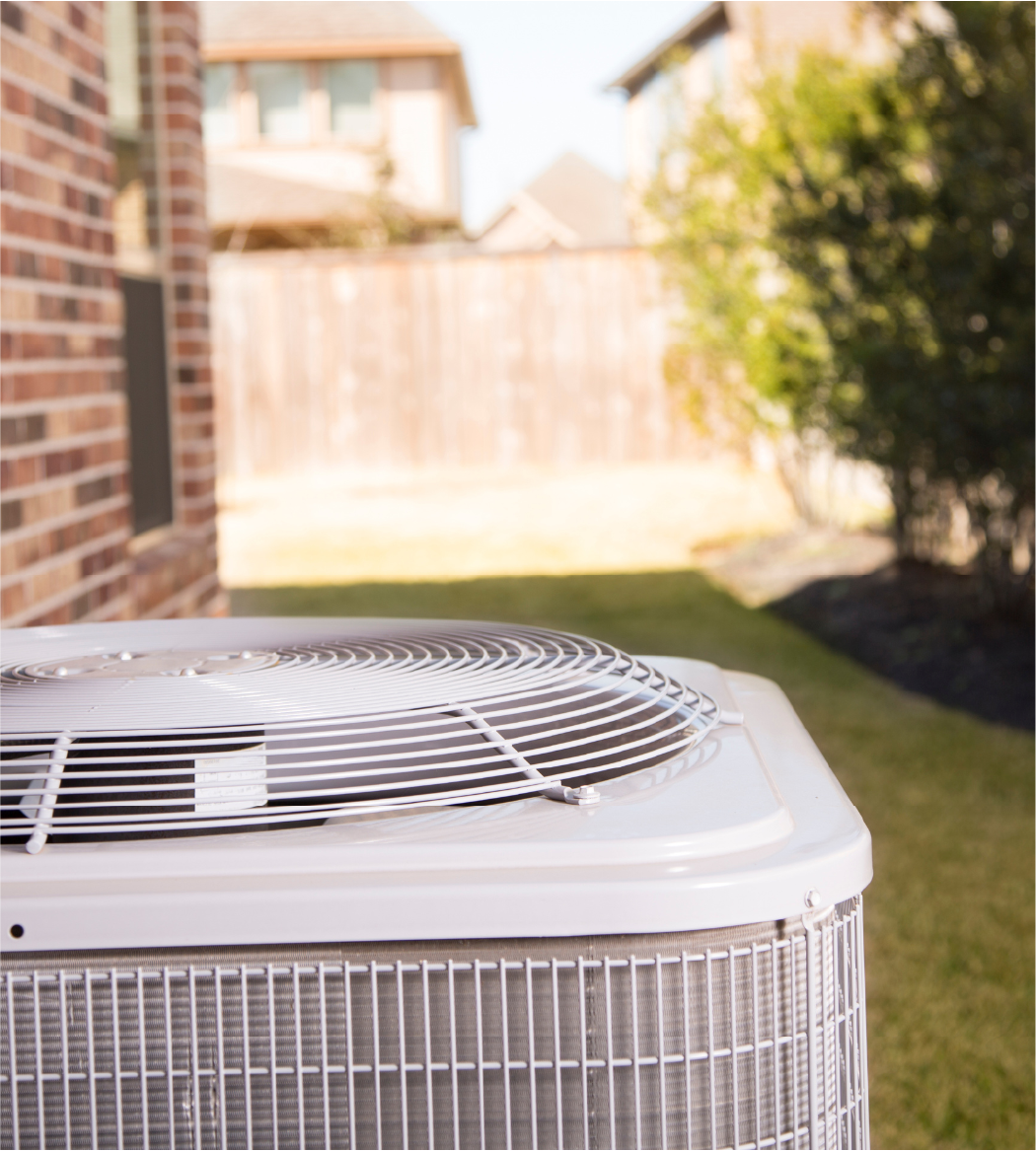 Outdoor air conditioning unit next to a brick house in a backyard with grass and shrubs.