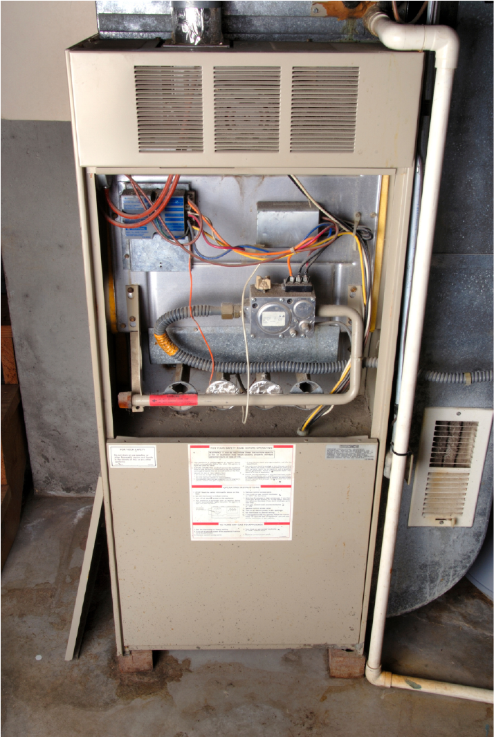 Old beige furnace unit in a basement with visible wiring, gas valve, and ventilation pipes.