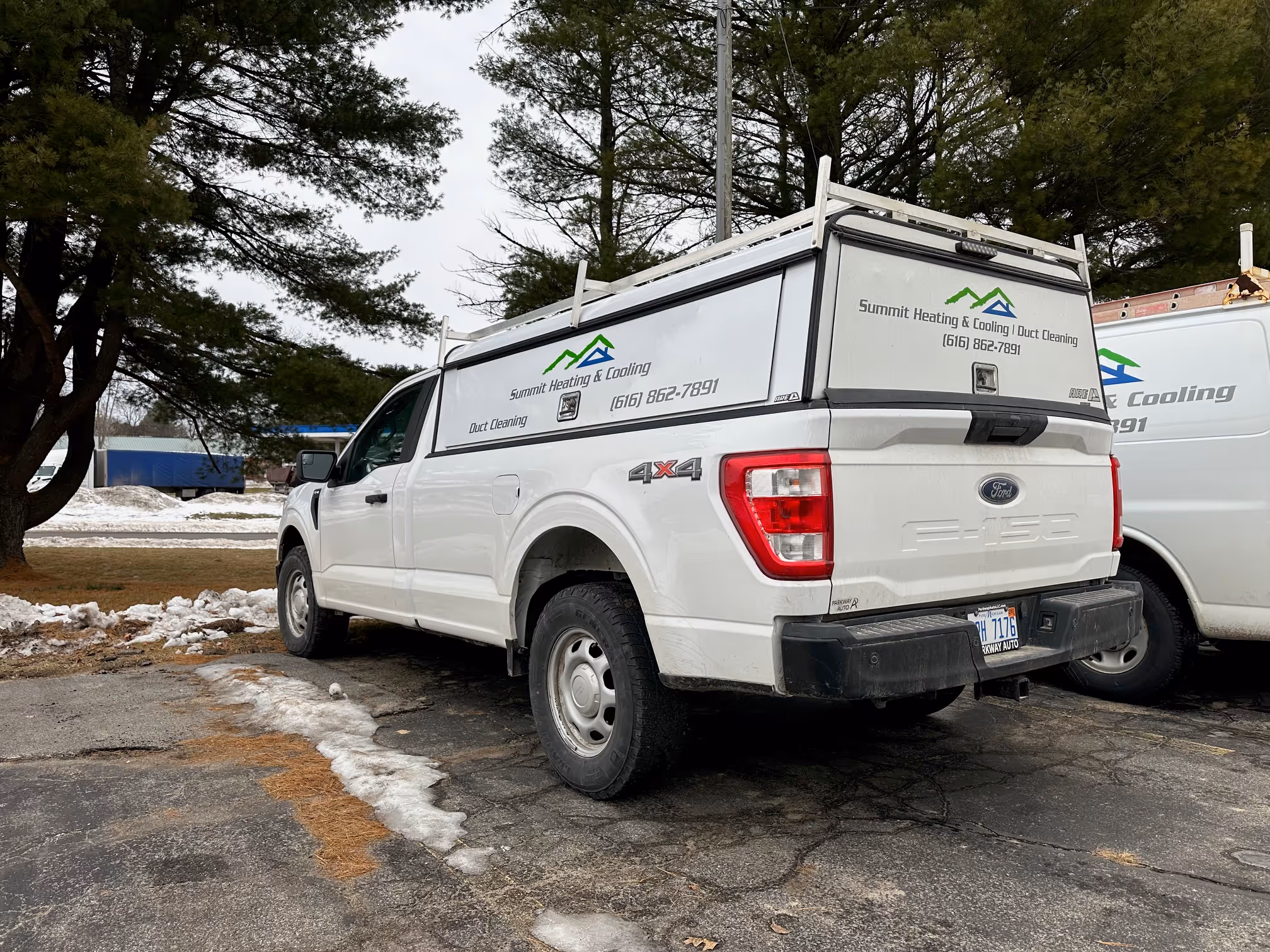 White Ford F-150 pickup truck with Summit Heating & Cooling and Duct Cleaning signage parked on a cracked asphalt surface with patches of snow and trees in the background.