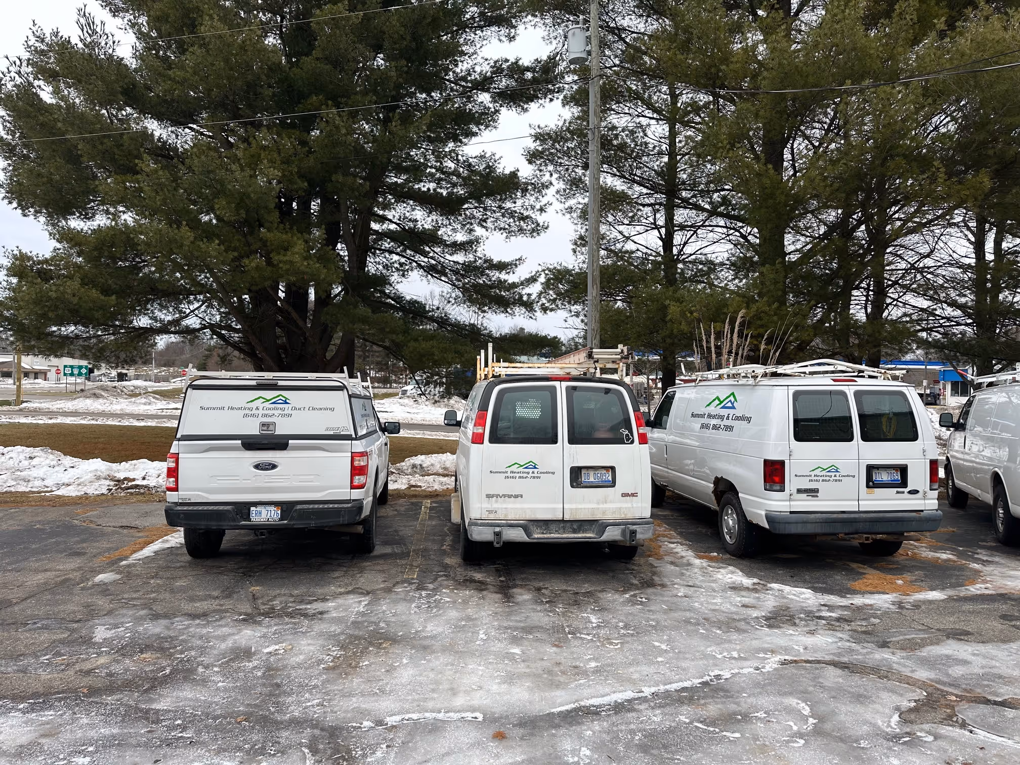 Three white service vehicles parked on a snowy asphalt lot under tall pine trees, each labeled Summit Heating & Cooling with a phone number.