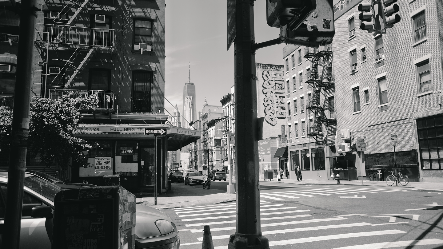 Black and white urban street scene with crosswalk, parked cars, fire escapes on buildings, and One World Trade Center in the background.