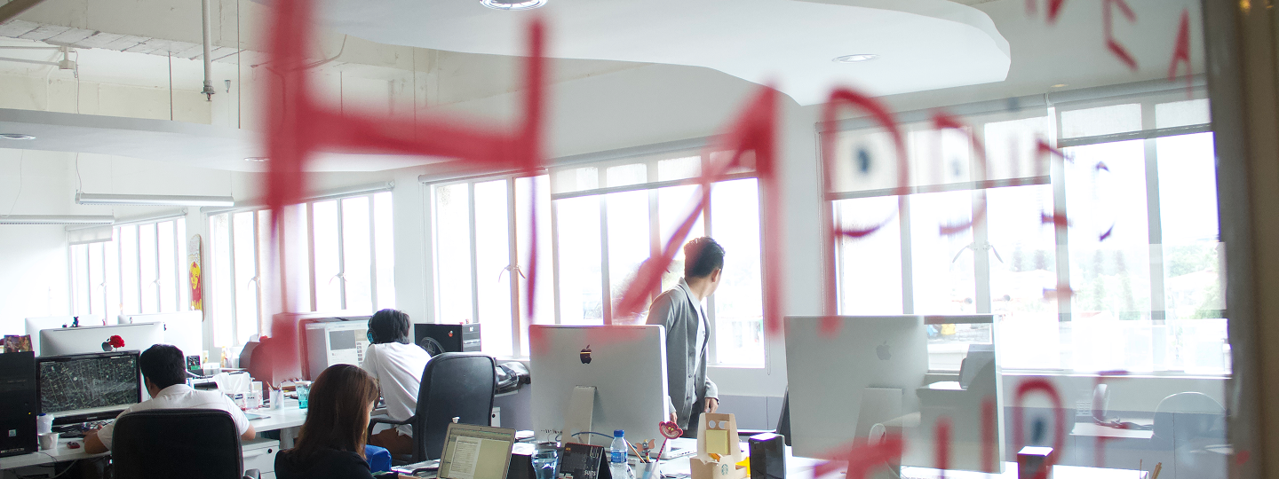 Office workspace with multiple people working at desks equipped with computers and laptops, viewed through glass with red writing.