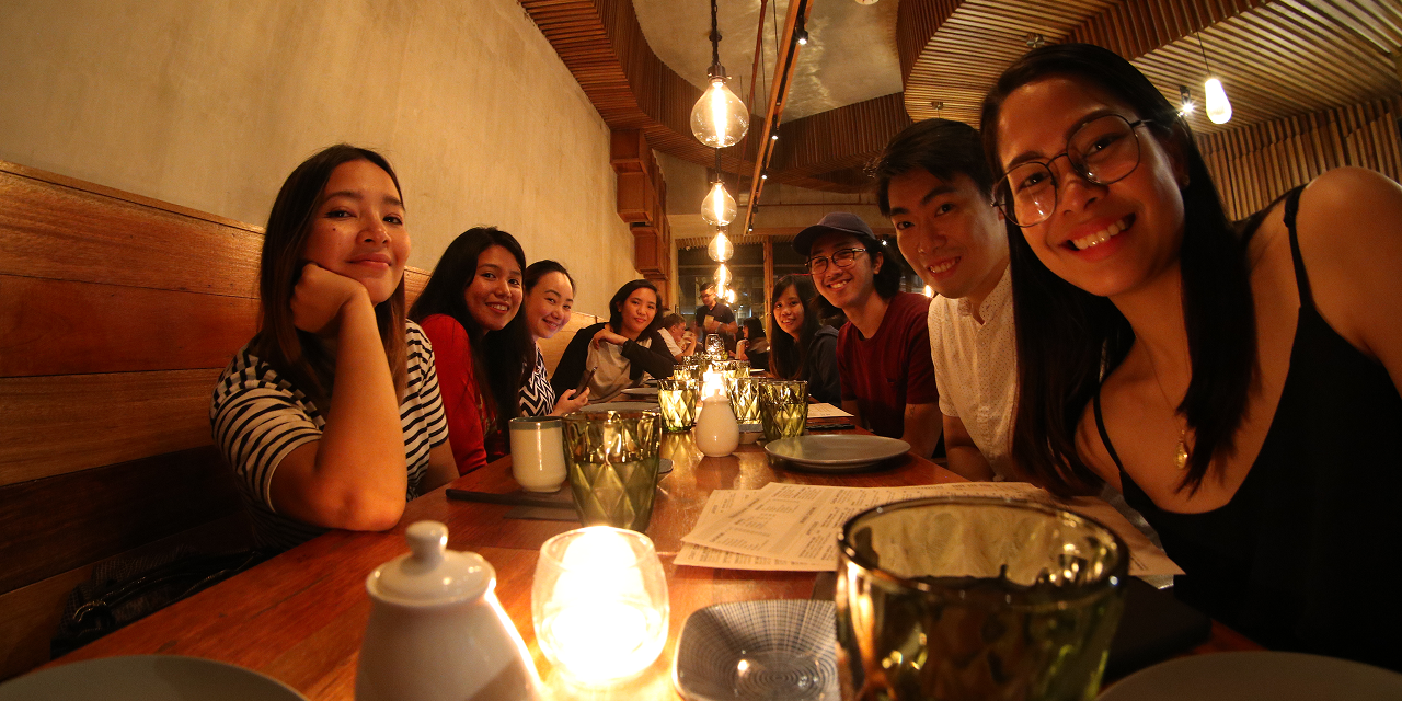Group of eight young adults smiling and sitting around a wooden table in a warmly lit restaurant.