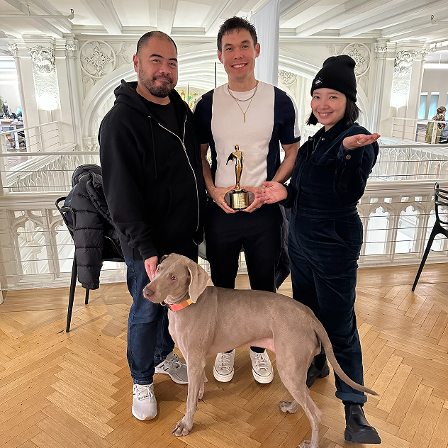 Three people standing indoors in front of a decorative railing, with the person in the center holding a trophy and a dog standing in front of them.