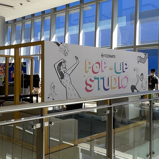 Indoor pop-up studio setup with large colorful sign reading 'POP-UP STUDIO' featuring illustrated people, inside a building with large windows and a person wearing a mask nearby.