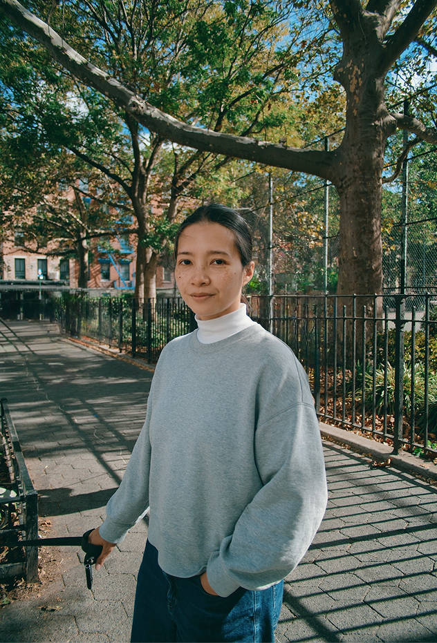 Young woman with dark hair in a gray sweatshirt and white turtleneck standing on a tree-lined city sidewalk on a sunny day.