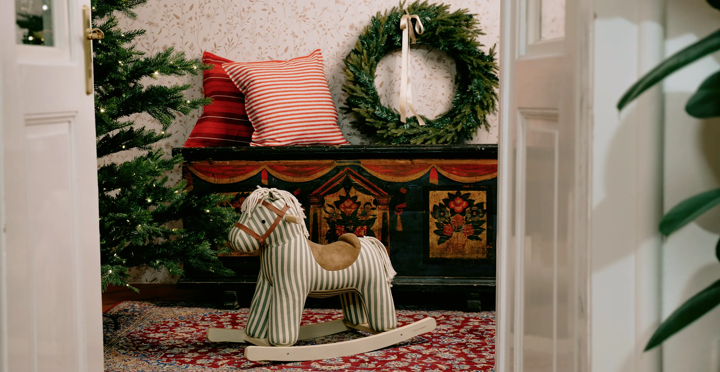 Striped fabric rocking horse on a patterned rug next to a decorated Christmas tree, red pillows, and a green wreath on a black chest.