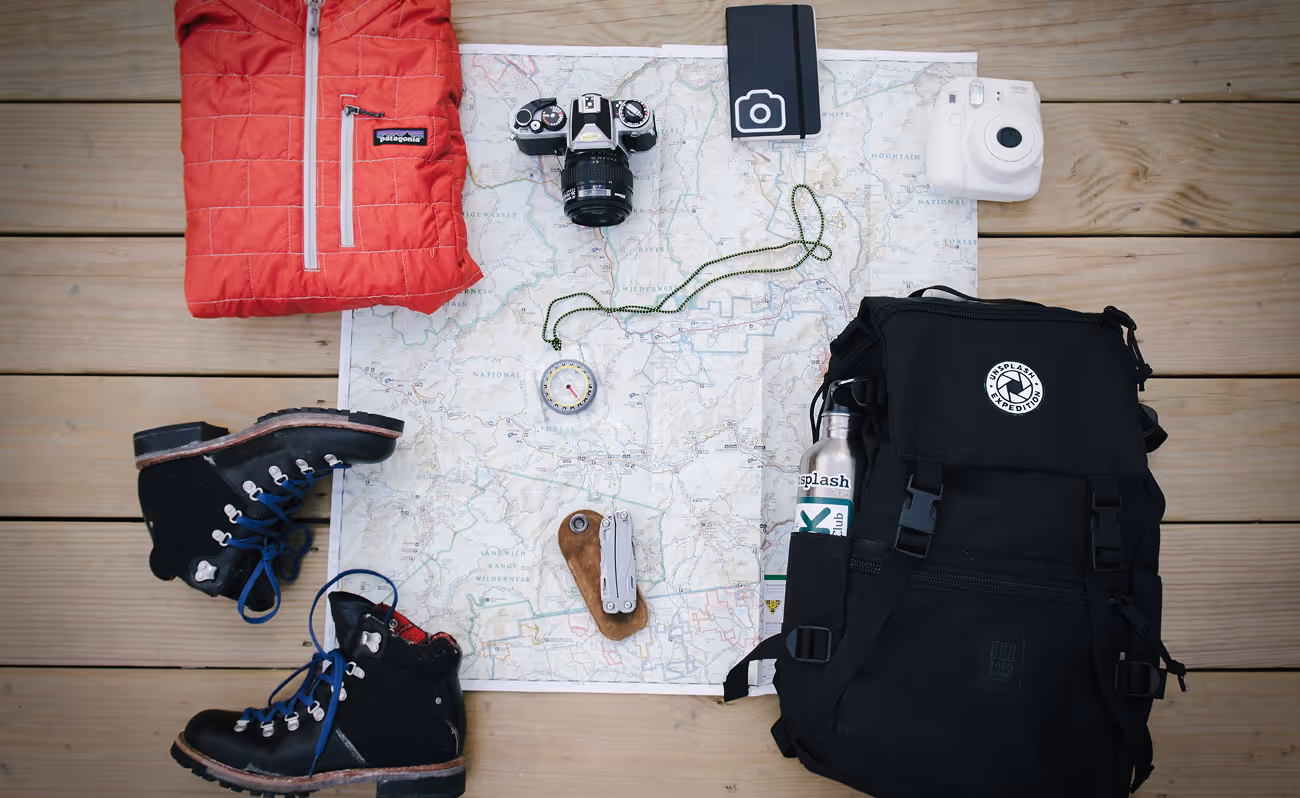 Top-down view of outdoor gear neatly arranged on a table, including a camera, boots, survival knife, and coat.