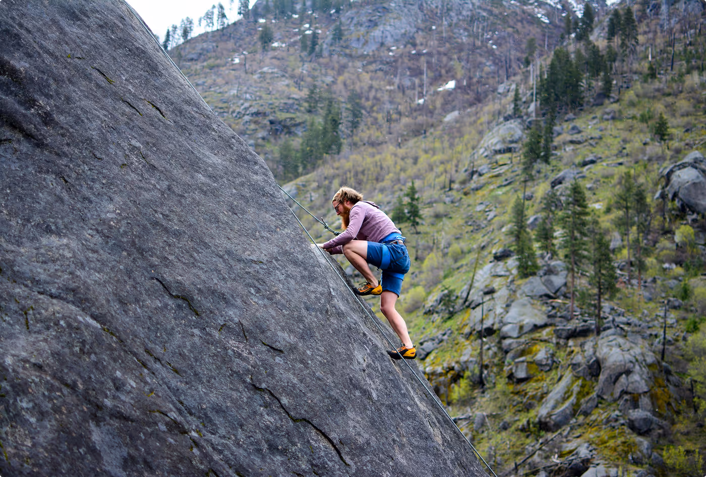 Person climbing a rock wall on a forested mountainside using ropes and a harness.