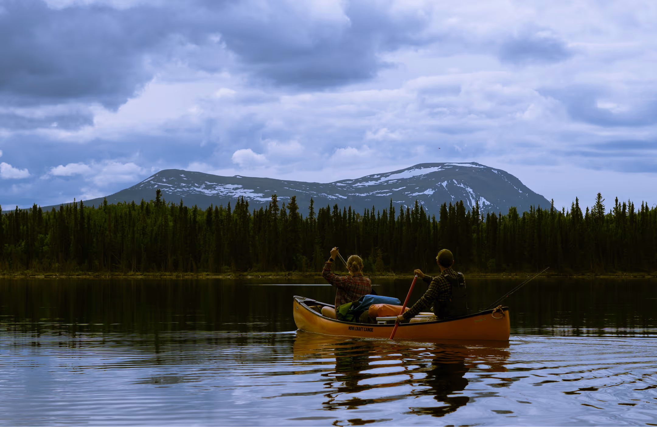 Two people paddling together in a canoe, looking toward a tree-lined lakeside with a mountain beyond.