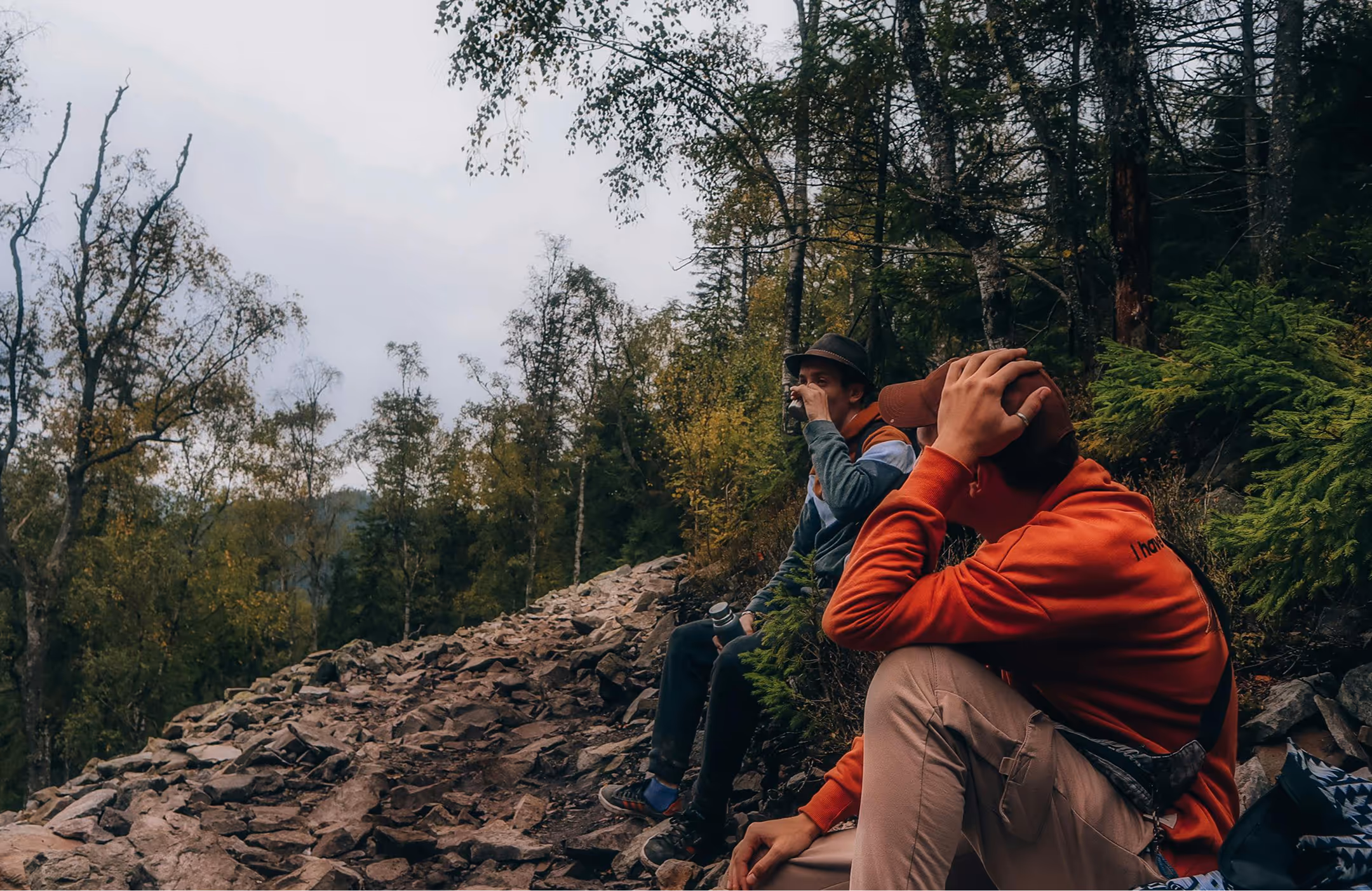 Two hikers sitting on a hillside during a break, with one drinking from a cup.
