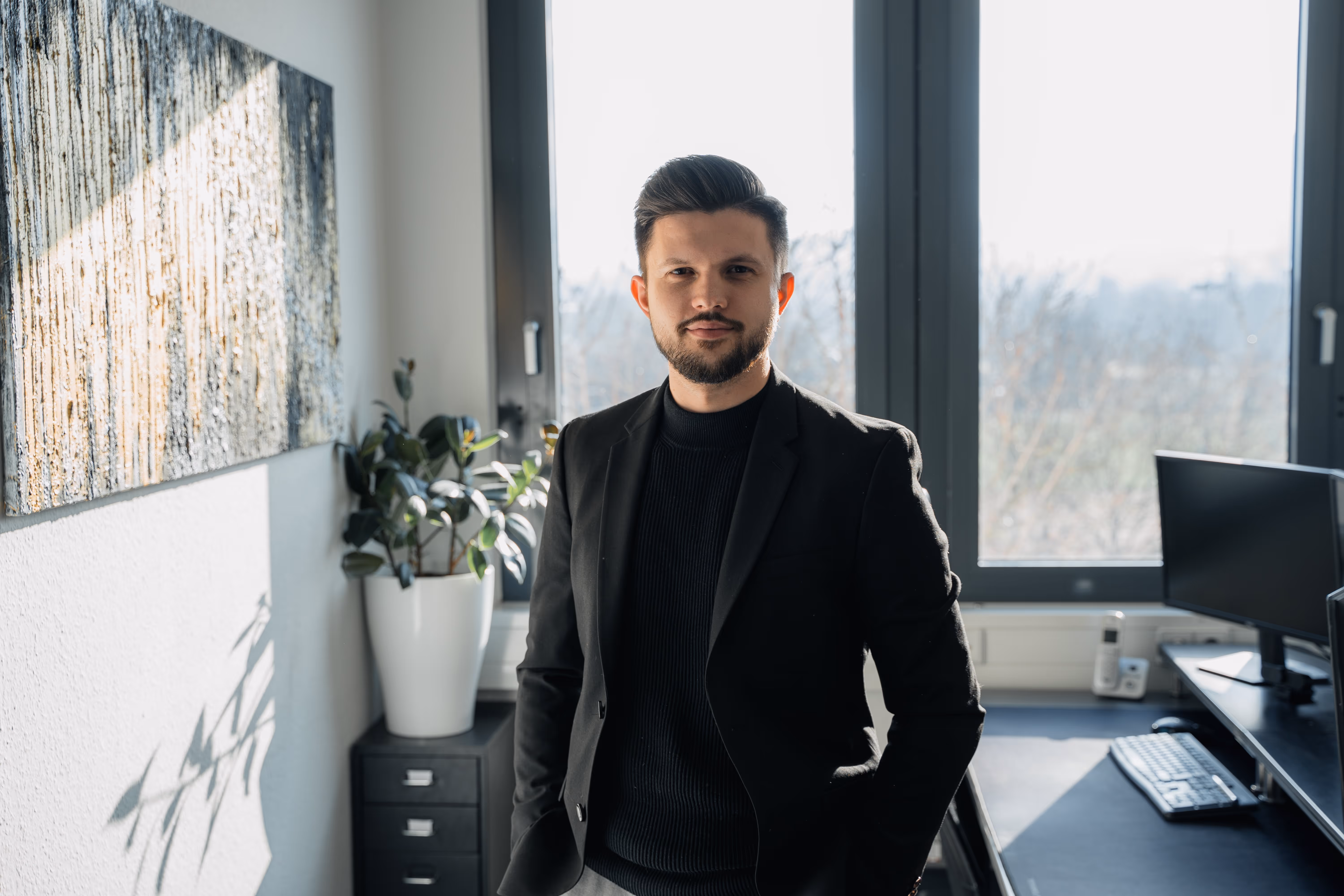 Man with beard and styled hair wearing a black blazer and sweater standing in a bright office with a plant, abstract painting, and computer.