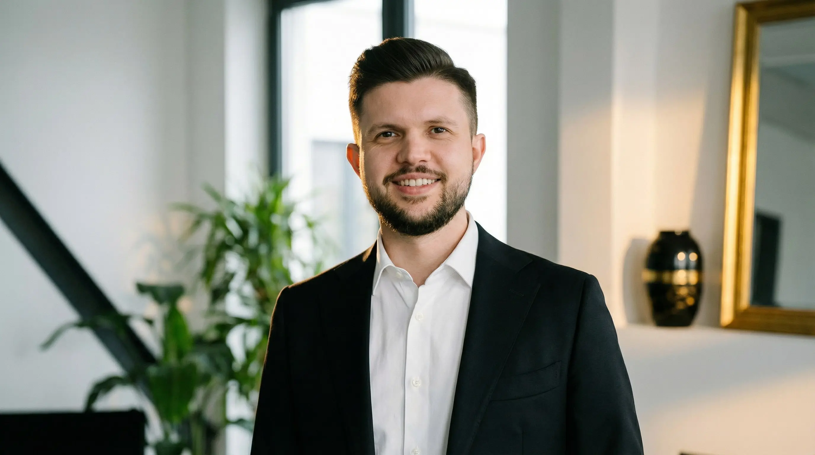 Smiling man with dark hair and beard wearing a black suit jacket and white shirt in a modern office setting.