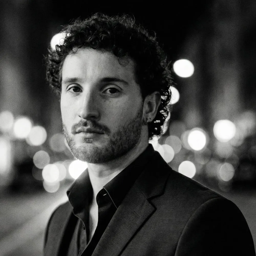 Black and white close-up of a man with curly hair and a beard wearing a dark suit, with blurred city lights in the background.