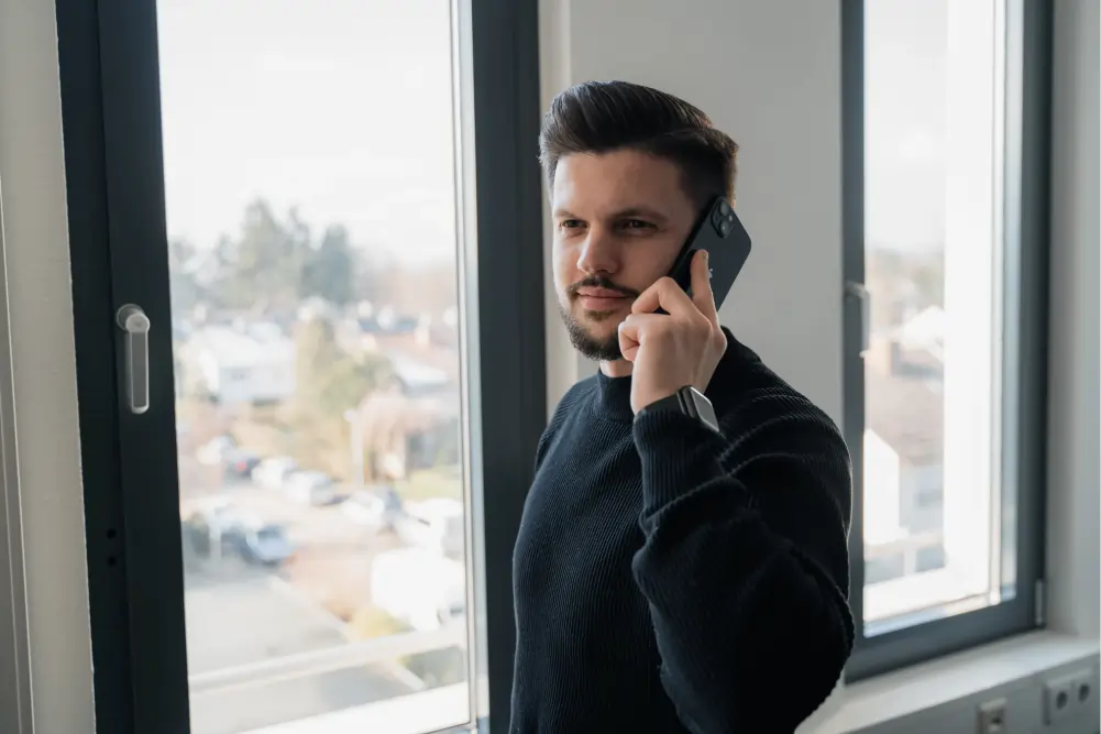 Man in a black sweater standing by a window talking on a smartphone.