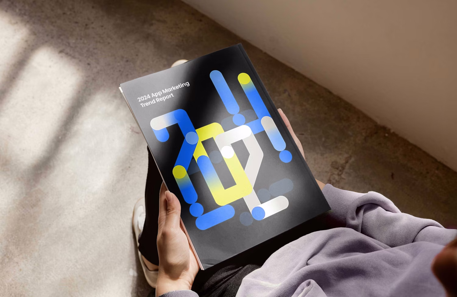 Person holding a black report titled '2024 App Marketing Trend Report' with blue, yellow, and white modern abstract design on the cover.