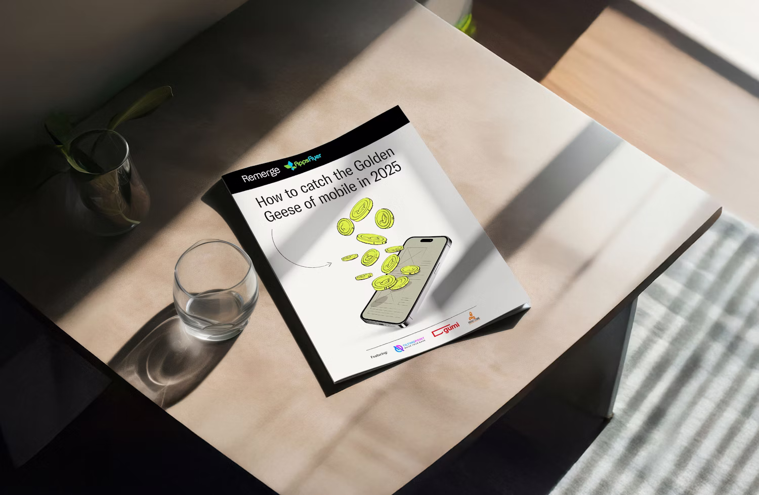 A booklet titled 'How to catch the Golden Geese of mobile in 2025' lying on a table next to a glass and a small plant pot.