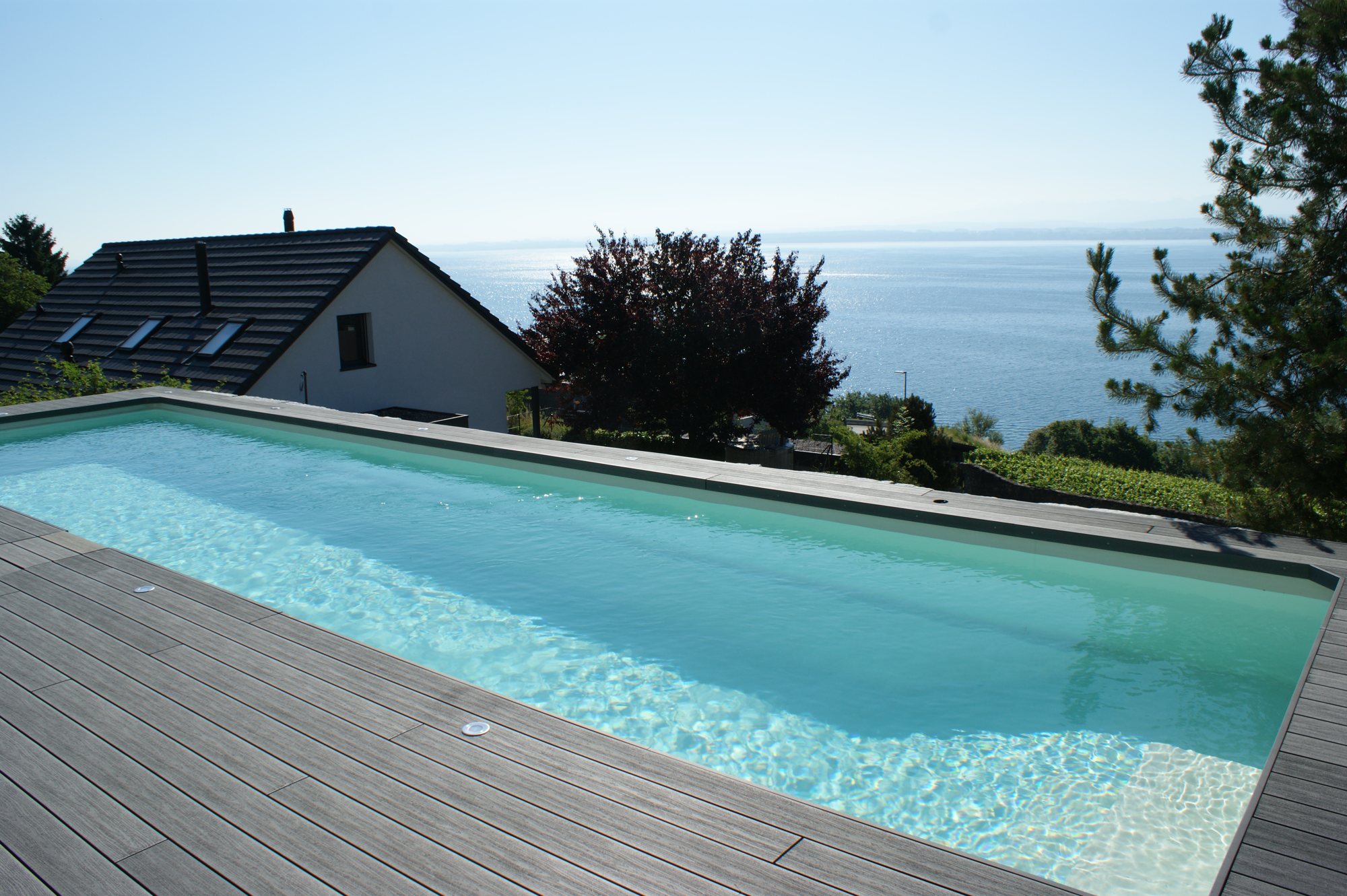 Piscine construite par Espace Piscines et Spas, avec terrasse en bois et vue sur le lac de Neuchâtel.