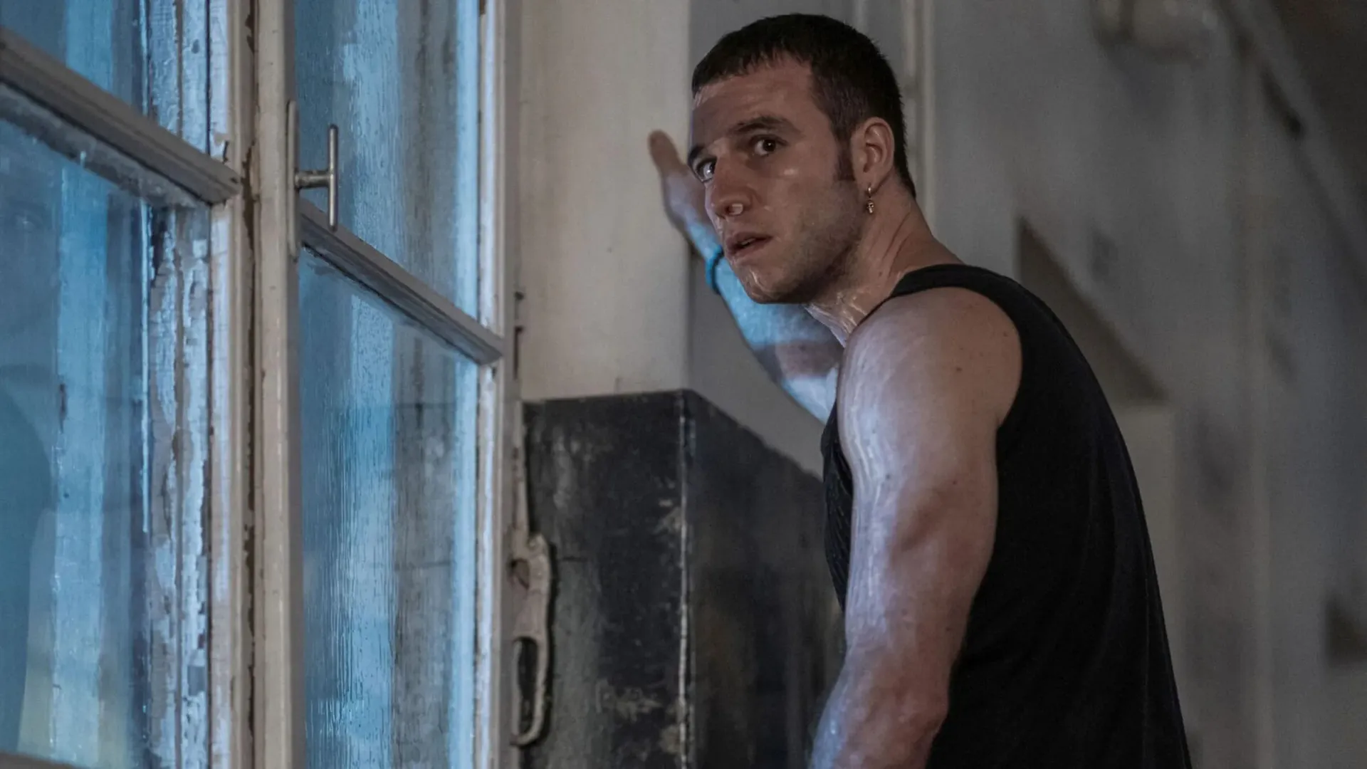 A man looking at the camera with a desperate expression in fron of a dirty window covered in sweat.