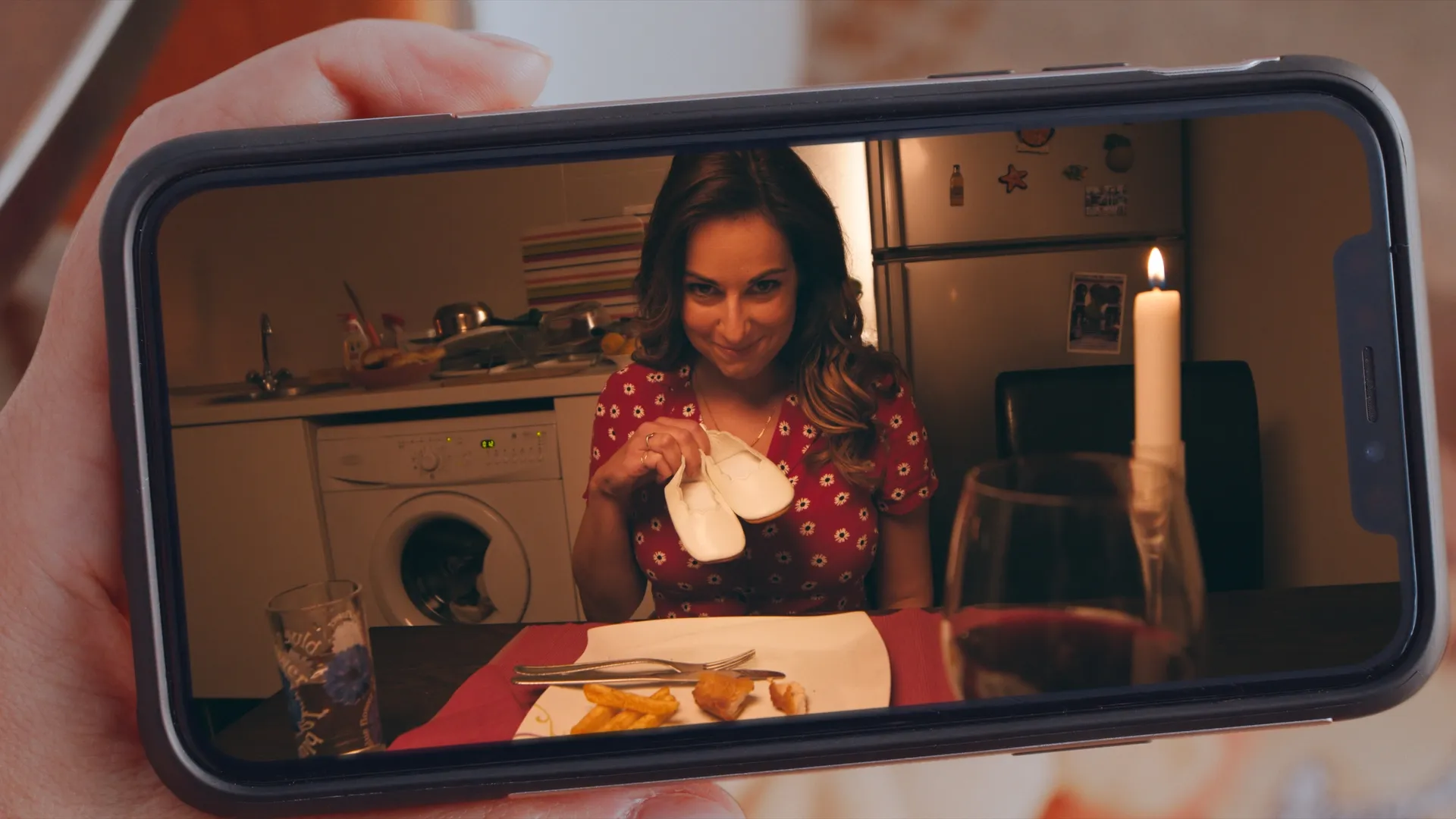 A person holding a phone showing a woman at a dinner table smiling and holding baby shoes.