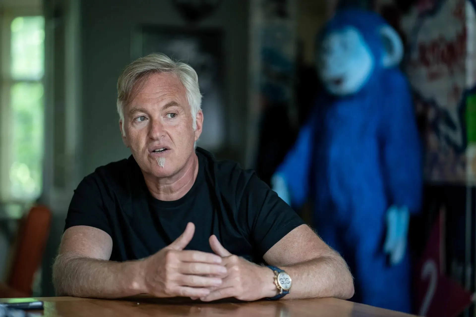 A gray haired man sitting at a desk explainingly gesturing with a costume of a blue monkey in the background.